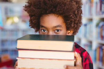 Portrait of african american schoolboy carrying stack of books in school library