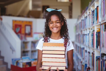 Portrait of smiling african american schoolgirl carrying stack of books in school library - obrazy, fototapety, plakaty