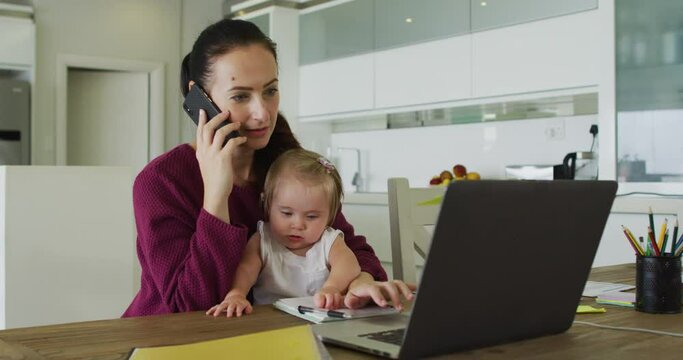 Caucasian mother holding her baby talking on smartphone and using laptop while working from home