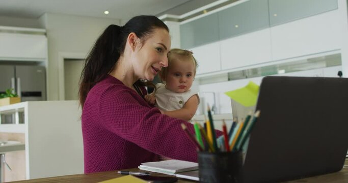 Caucasian mother holding her baby using laptop while working from home