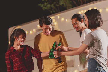 Group of young diverse friends holding the bottle drinking talking and enjoying a picnic in the park in the evening playing music having fun on the camping area by the nature.