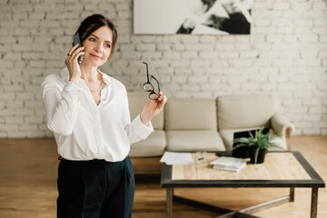 Middle aged business woman working at home. Talking on the phone and and smiling, holding eyeglasses.