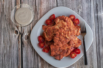 A square white plate with breaded chicken boneless legs and cherry tomatos and a fork, over a rustic table  with a glass cup of dressing or garvy, flat lay