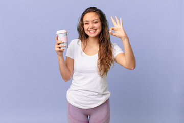 young pretty woman feeling happy, showing approval with okay gesture and holding a coffee