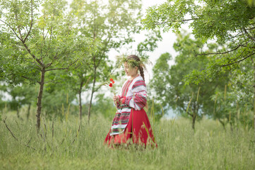 A girl in a Belarusian folk costume stands in the forest with a wreath on her head and flowers in her hands. The concept of the traditions of the Slavic people.