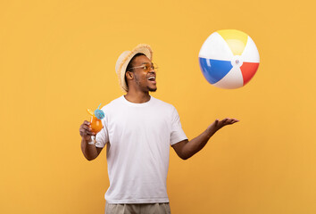 Happy black man drinking tropical fruit cocktail and playing with inflatable beach ball on yellow...