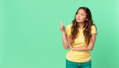 young pretty woman shrugging, feeling confused and uncertain and pointing to the copy space