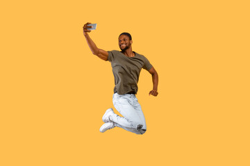 Fototapeta na wymiar Positive african american man jumping and making selfie on smartphone over yellow background