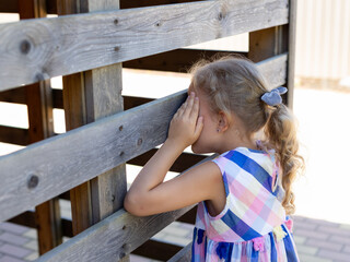 cute little girl covering her eyes with hands, playing hide and seek standing near wooden fence. Outdoor activities with kids. Childhood