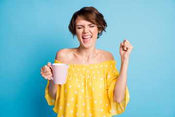Photo portrait of excited girl celebrating with raised fist holding cup in one hand isolated on vivid blue colored background