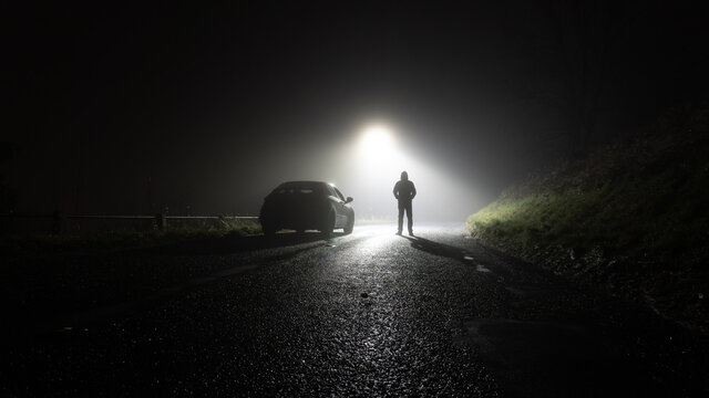 A lone car, parked on the side of the road, underneath a street light, with a hooded figure, on a spooky, scary, rural, country road. On a foggy winters night