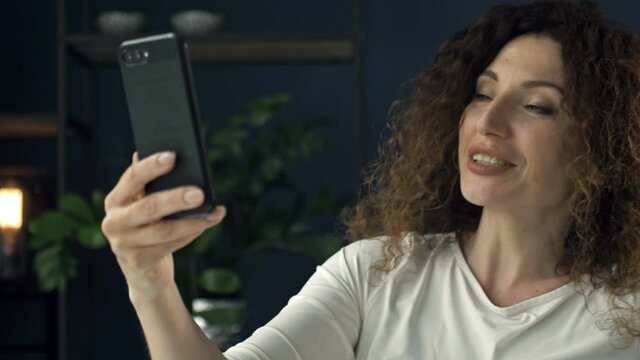 Beautiful 40 year old woman with a mobile phone in her hand waves and smiles happily during a video call. She looks like she's flirting with someone.