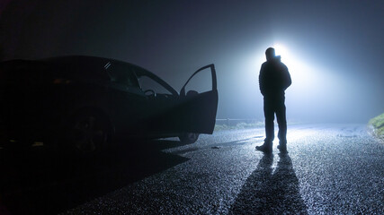 A man standing next to a car, with door open, parked on the side of the road, underneath a street light, on a spooky, scary, rural, country road. On a foggy winters night - Powered by Adobe