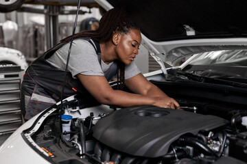 Fototapeta na wymiar african woman under the hood of white car. woman in uniform mechanic repairing a car in car service. portrait of young black female enjoying work with automobile, vehicle