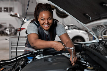 Fototapeta na wymiar African Female Checks an Engine Breakdown. Car Service Employee Woman Fix the Engine Component. Modern Clean Workshop. Black Woman In Overalls Looks At Camera Smiling