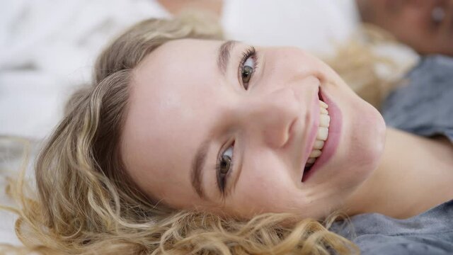 Headshot portrait of cheerful slim gorgeous Caucasian blond woman looking at camera lying in cozy bed at home. Close-up face of happy millennial enjoying weekend morning in bedroom indoors