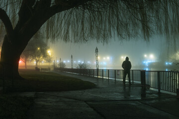 A mysterious moody hooded figure silhouetted against street lights by a river on a foggy...