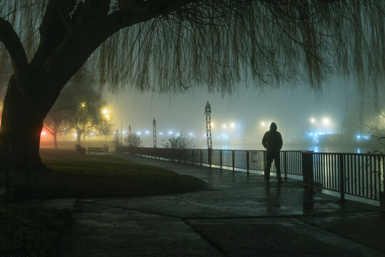 A mysterious moody hooded figure silhouetted against street lights by a river on a foggy atmospheric winters night