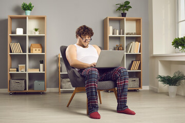 Funny serious fat man in pajamas and eyewear looking at screen working online on laptop from home...
