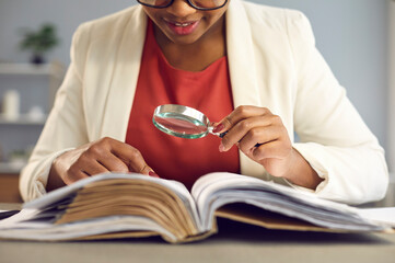 Young woman sitting at desk, holding magnifying glass and reading book. Businesswoman, business...
