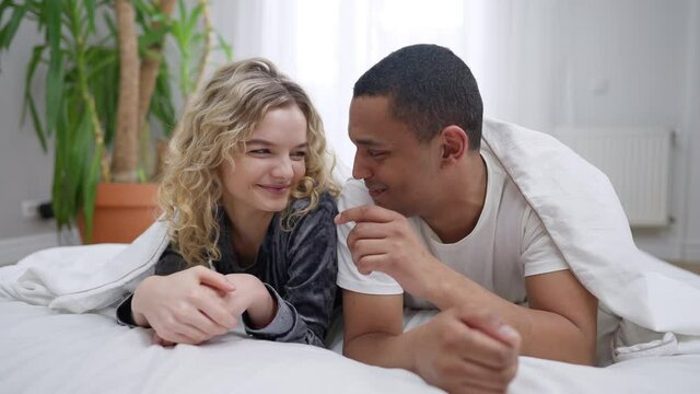 Shy millennial interracial couple looking at each other smiling lying in bed under blanket. Positive happy lovers enjoying morning in bedroom. African American man touching nose of Caucasian woman