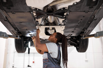 Side view on black female auto mechanic fixing car, mechanic repairing car on lift in garage. pretty afro american lady in overalls concentrated on work, repair using instruments tools