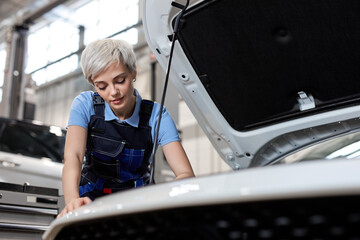 Woman auto mechanic in uniform examine car engine breakdown problem in front of automotive vehicle car hood. Safety technical inspection care check service maintenance