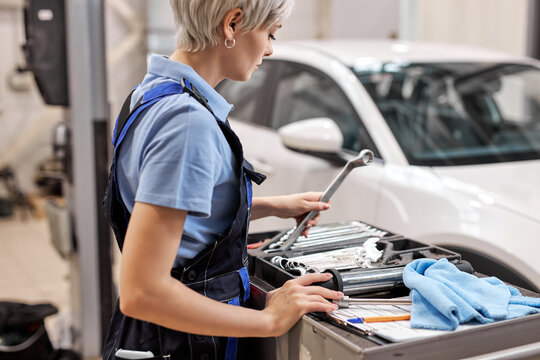 Portrait of young female auto mechanic taking tool instruments for car repairing, having blue rag, box with many various instruments, woman in overalls uniform is ready for work in garage
