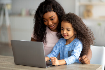 Home schooling during covid concept. Cheerful black mom helping her daughter with online homework on laptop pc