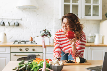 Gorgeous young woman plus size body positive in red longsleeve records cooking process on phone camera at home kitchen. Smiling woman food blogger do video stream on mobile on tripod