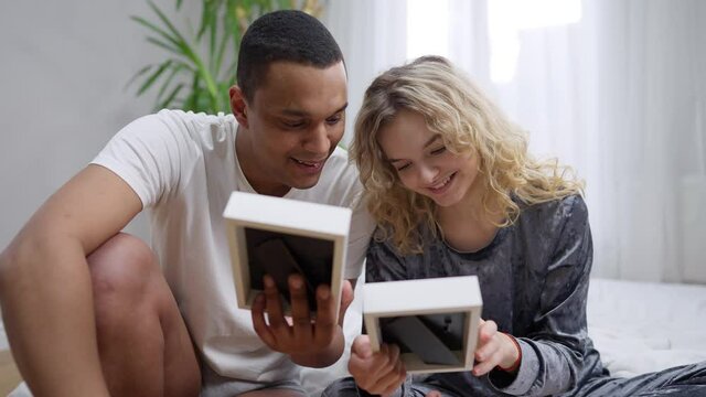 Happy young couple talking smiling looking at pictures sitting in bedroom in the morning. Portrait of joyful Caucasian woman and African American man recolling memories of love indoors at home