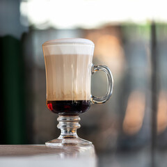 Three-layer coffee with foam in tall glass cup with handle on table against of blurred background. Side view.