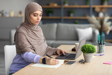 Muslim woman sitting at desk, using computer writing in notebook