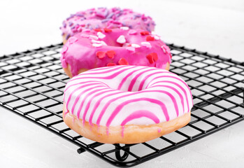 Three pink donuts with different icings and sprinkles on pastry rack on the white table. American...
