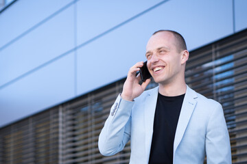 Bottom view perspective of young short-haired businessman in a blue suit standing in front of a corporate building solves a business phone call.