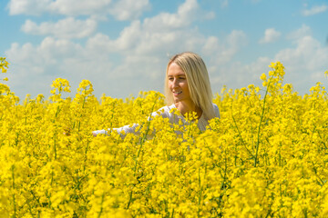 Naklejka na ściany i meble Smiling young blonde woman with her arm outstretched in a white shirt in a rapeseed field touching flowers on a sunny day