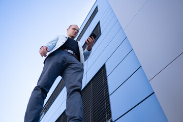 Bottom view perspective of young short-haired businessman in a blue suit standing in front of a corporate building solves a business phone call.