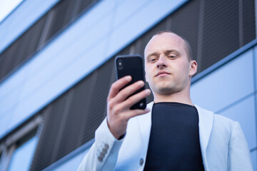 Bottom view perspective of young short-haired businessman in a blue suit standing in front of a corporate building and writes a text message on mobile.