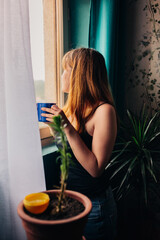 Young woman with a cup standing near the window enjoying relaxing in her home