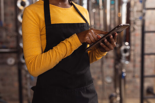 Mid section of african american female worker wearing apron using digital tablet at gin distillery