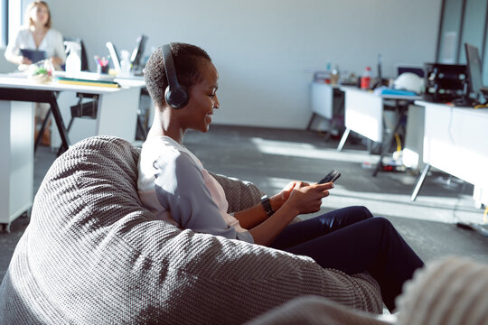 Smiling african american businesswoman sitting in armchair, wearing headphones, using smartphone