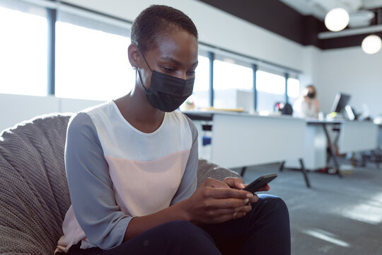 Smiling african american businesswoman sitting in armchair, wearing face mask, using smartphone