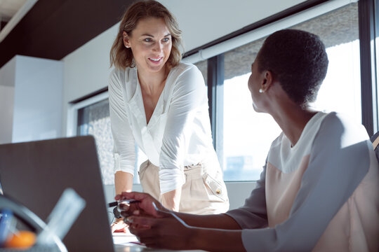 Two diverse smiling businesswomen working together, using laptop, talking