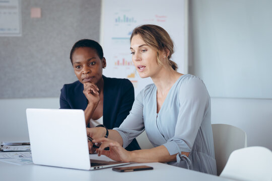 Two diverse businesswomen sitting at desk, using laptop, talking