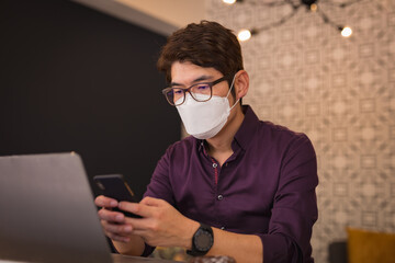Asian businessman wearing face mask using smartphone and laptop in hotel lobby
