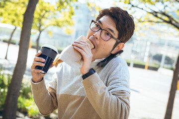 Asian businessman using eating sandwich and holding takeaway coffee sitting in city street