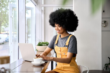 Young happy smiling black hipster girl of 20s with afro hair sitting in cafe indoors working using laptop having online web videoconference, talking to friends. - obrazy, fototapety, plakaty