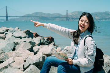 cheerful asian girl backpacker holding a map is smiling at camera while pointing at distance by sea...