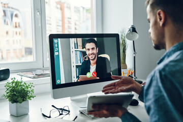 Serious young man talking to collegue by video call while sitting in office