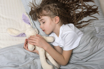A cute sleeping  little girl with a long wavy hair holding a white hare in her hands  lying on her...